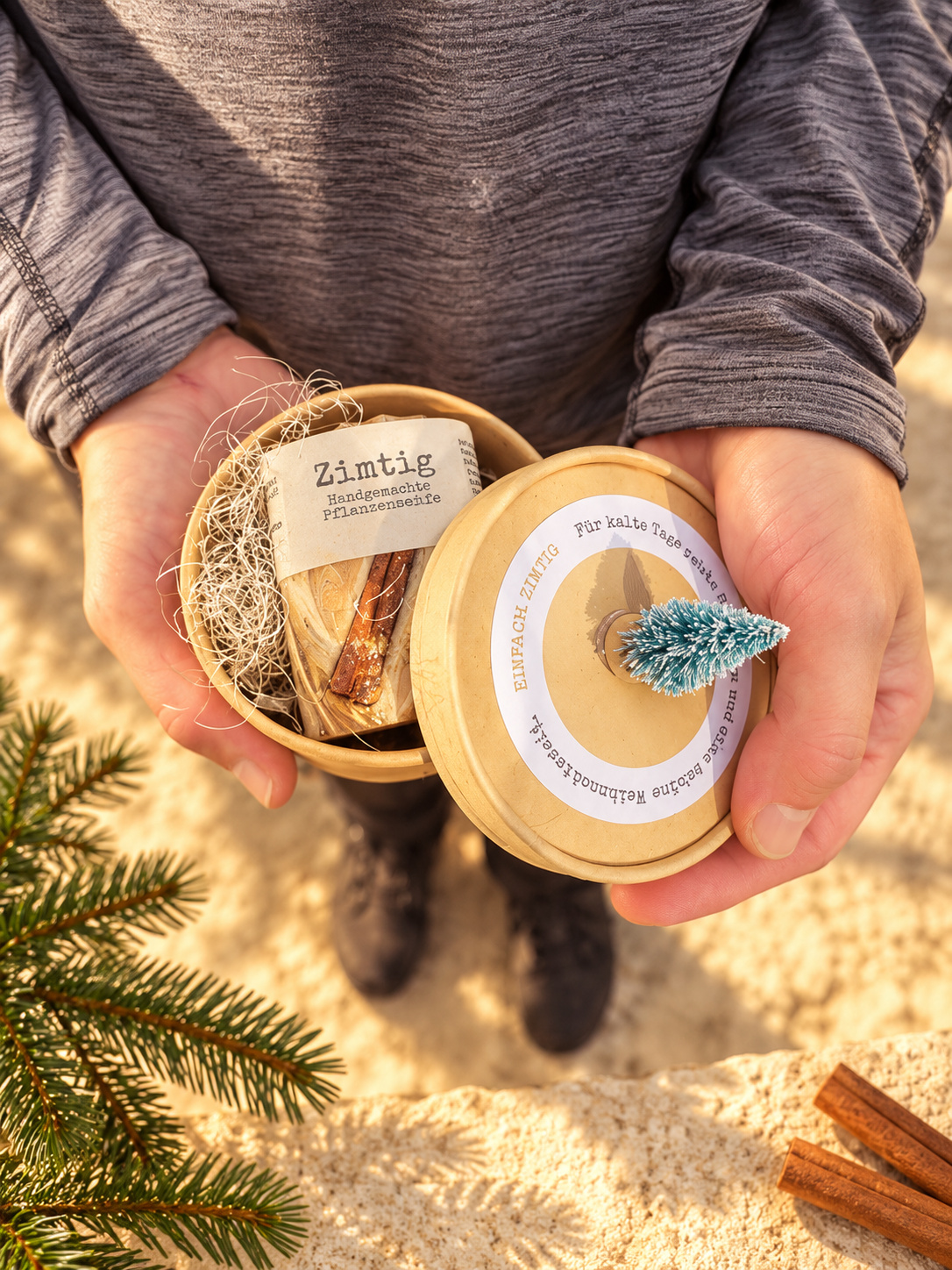 Zwei Hände halten die offene Geschenkbox „einfach Zimtig“ mit marmorierter Zimtseife und einem Deckel mit kleinem Tannenbäumchen im warmen Winterlicht.