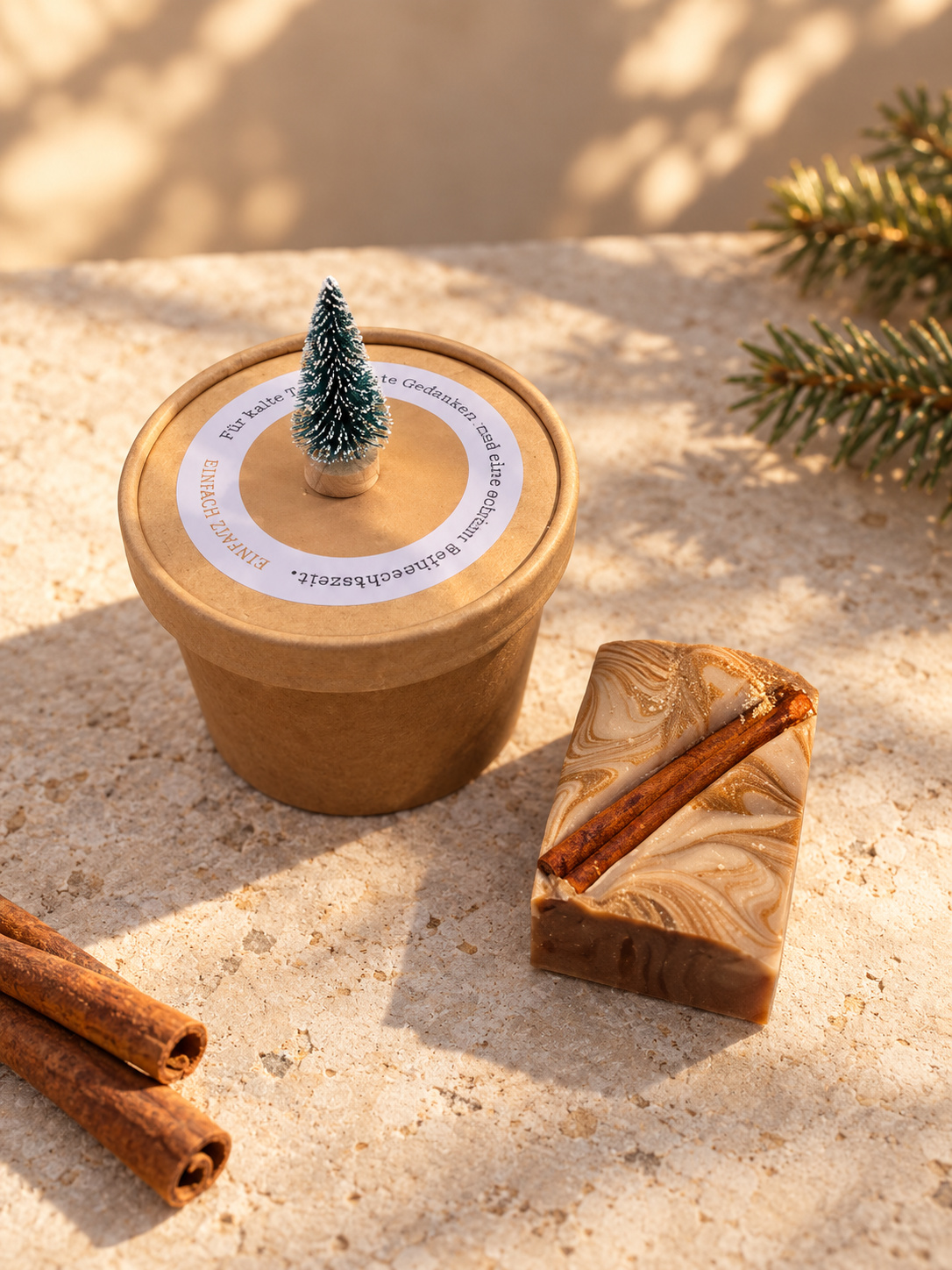 Geschlossene Geschenkbox „einfach Zimtig“ mit kleinem Tannenbäumchen neben einer liegenden, marmorierten Zimtseife auf hellem Stein im warmen Winterlicht.